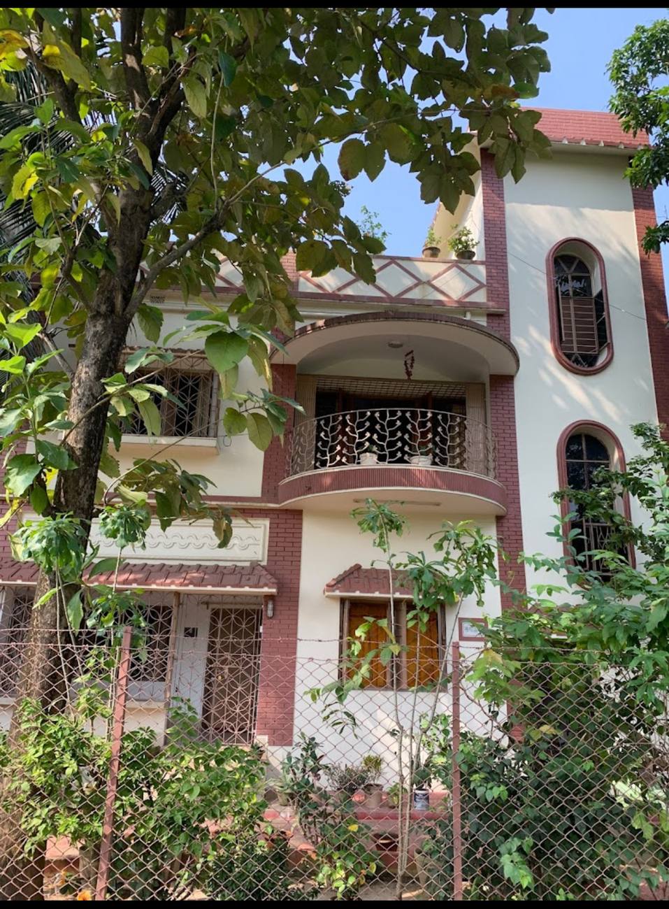 The front of Mitra Stay: a cream house with oxblood brick trim, arched windows and a curved balcony, seen through the leaves of a teak tree.