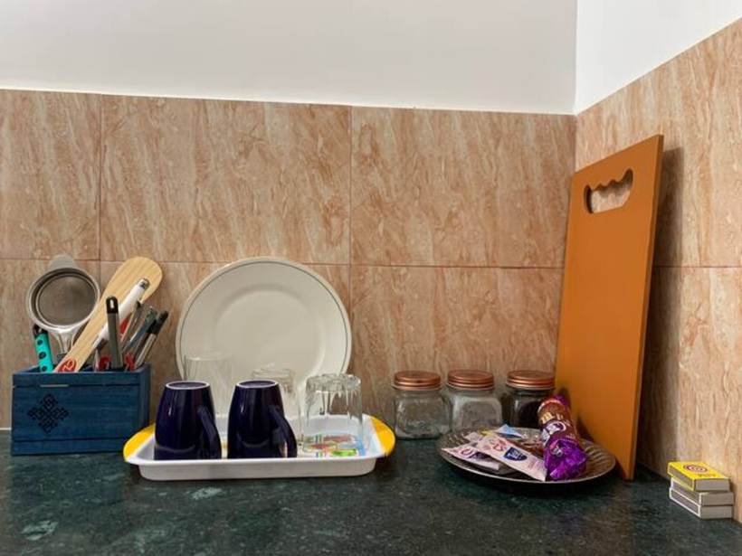 A kitchen counter with a cutlery caddy, plates, glasses, blue mugs on a tray, storage jars, an orange chopping board and a plate of wrapped sweets.
