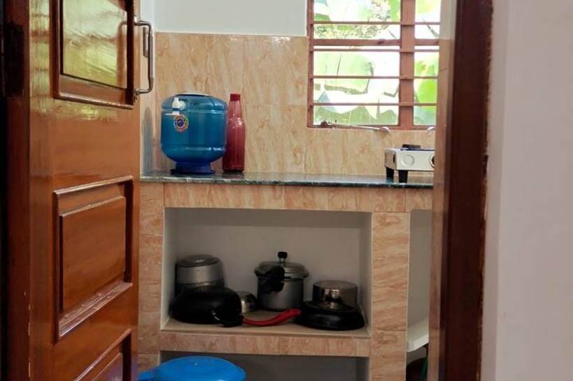 A small kitchen through an open wooden door: a blue water dispenser, a gas stove on a green marble counter, and pressure cookers and pans stacked in the shelf below.