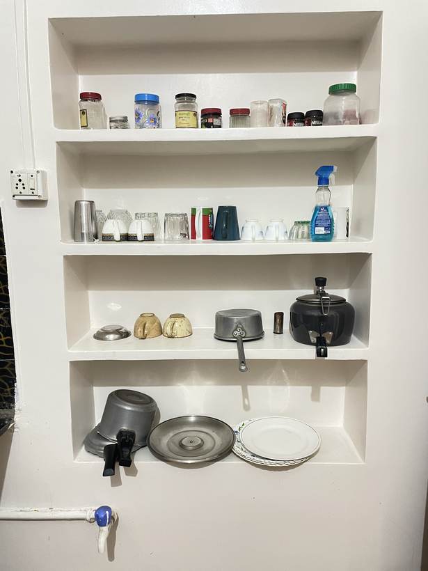 Four shelves recessed into a white wall holding storage jars, glasses, teacups, a pan and a black kettle.
