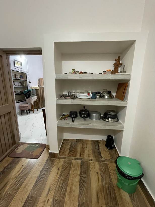 A tall recessed shelf unit of pots and jars beside an open door looking through into the bedroom with its bookshelf and television.