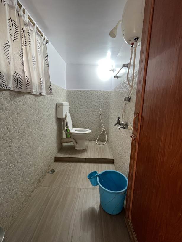 A washroom with pebble-textured white tiles, a toilet, a shower with an electric geyser above it, and a block-print curtain at the window.