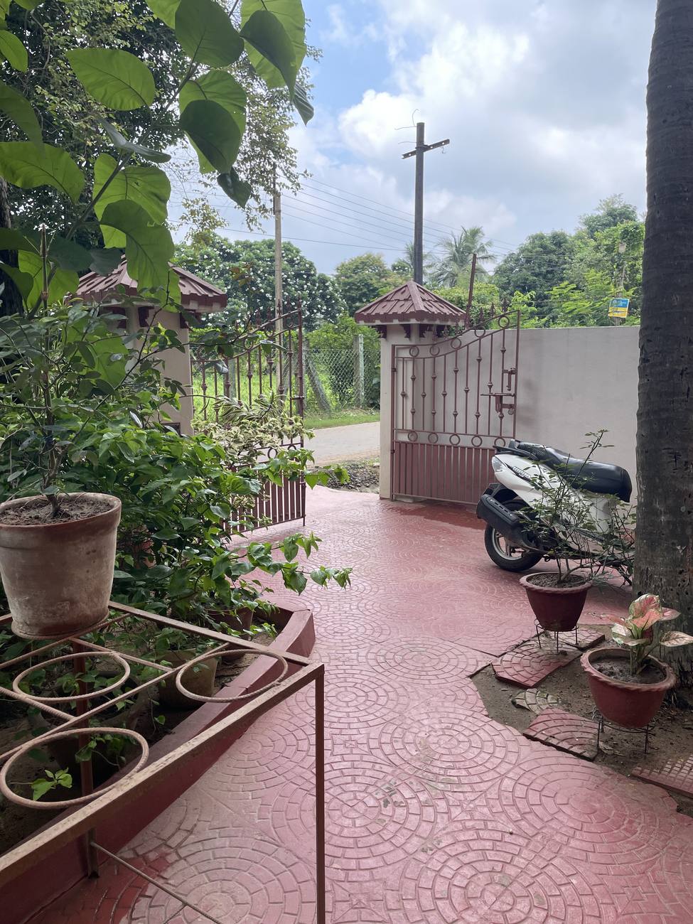 The gate, iron with tiled caps, opening onto a quiet road, with the red-oxide drive and a scooter parked inside.