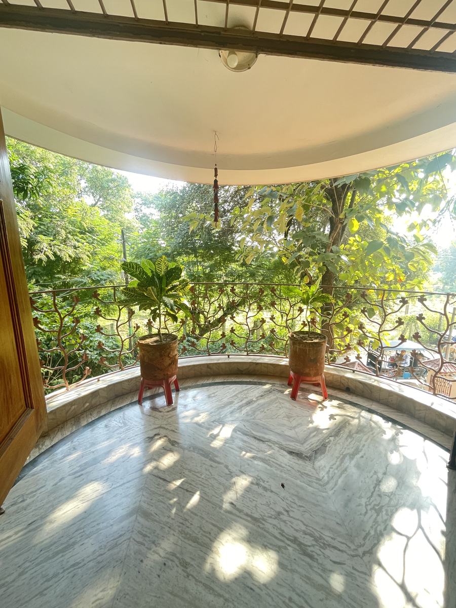 A curved balcony with a polished marble floor, a wrought-iron railing in a branching leaf pattern, two crotons in terracotta pots, and green trees filling the view beyond.