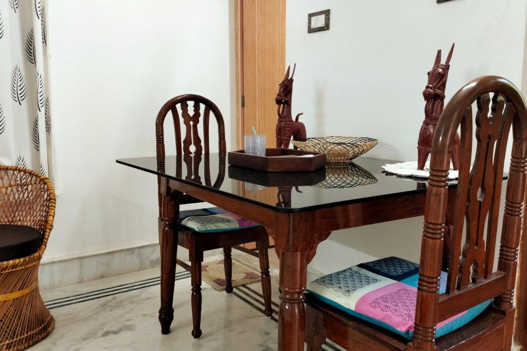 A glass-topped wooden dining table with carved chairs, block-print seat cushions, Bankura terracotta horses as a centrepiece, and a cane chair beside it.