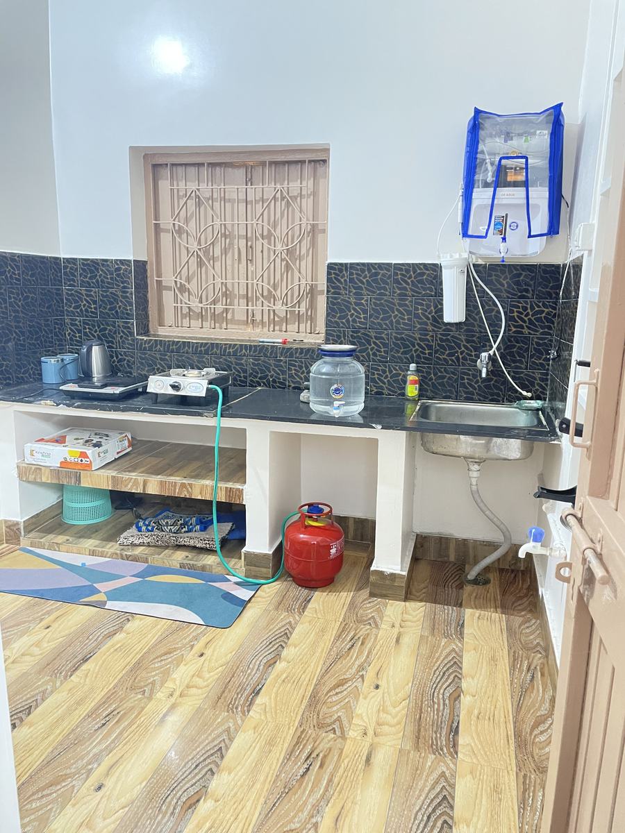 A kitchen with a black granite counter, a two-burner gas stove and cylinder, an electric kettle, a stainless sink, a wall-mounted water purifier, dark patterned tiles and a wood-look floor.