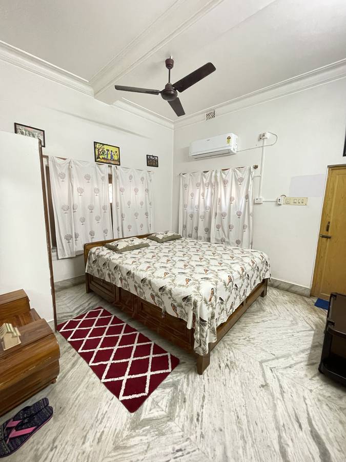 A bright bedroom with a wooden storage bed, bird-print bedcover, block-print curtains and an air conditioner.