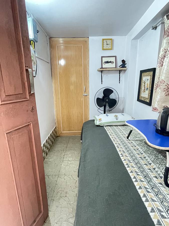 A small simple room with a green bedcover, a standing fan, a shelf, and a blue folding table holding an electric kettle.