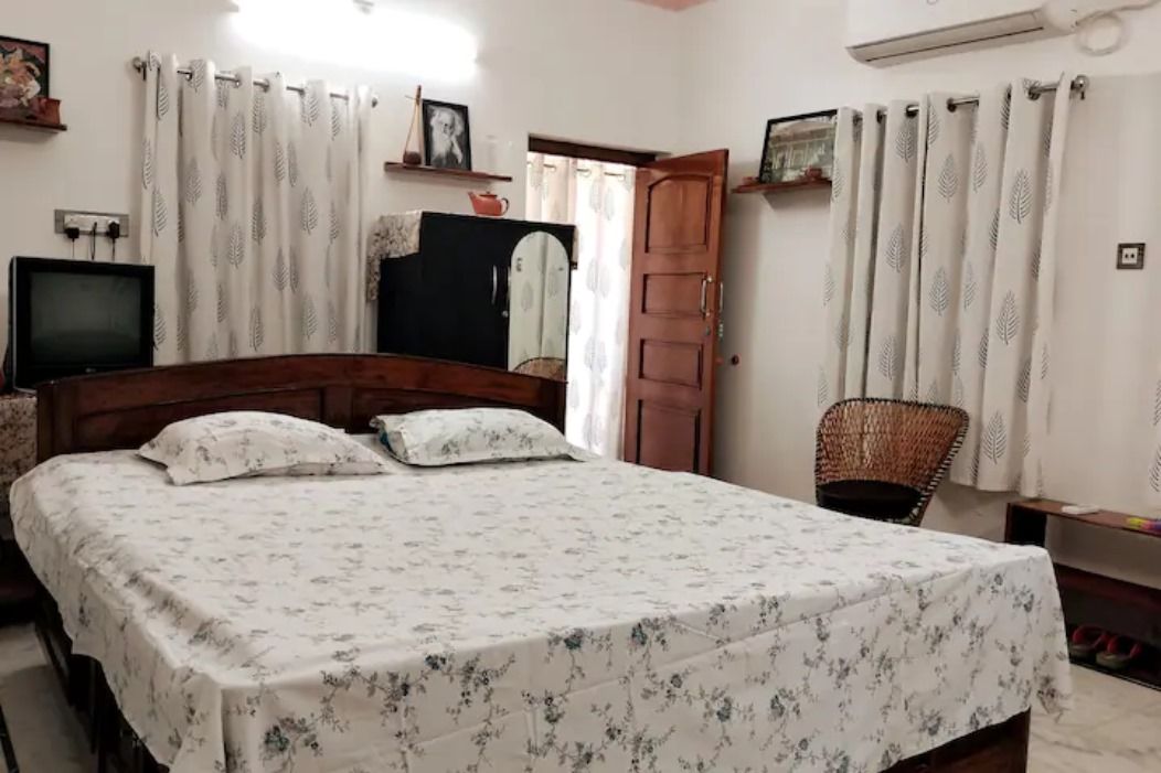 A bedroom with a carved wooden bed and floral bedcover, leaf-print curtains, framed photographs on narrow shelves, a cane chair, and a polished wooden door standing open to the balcony.