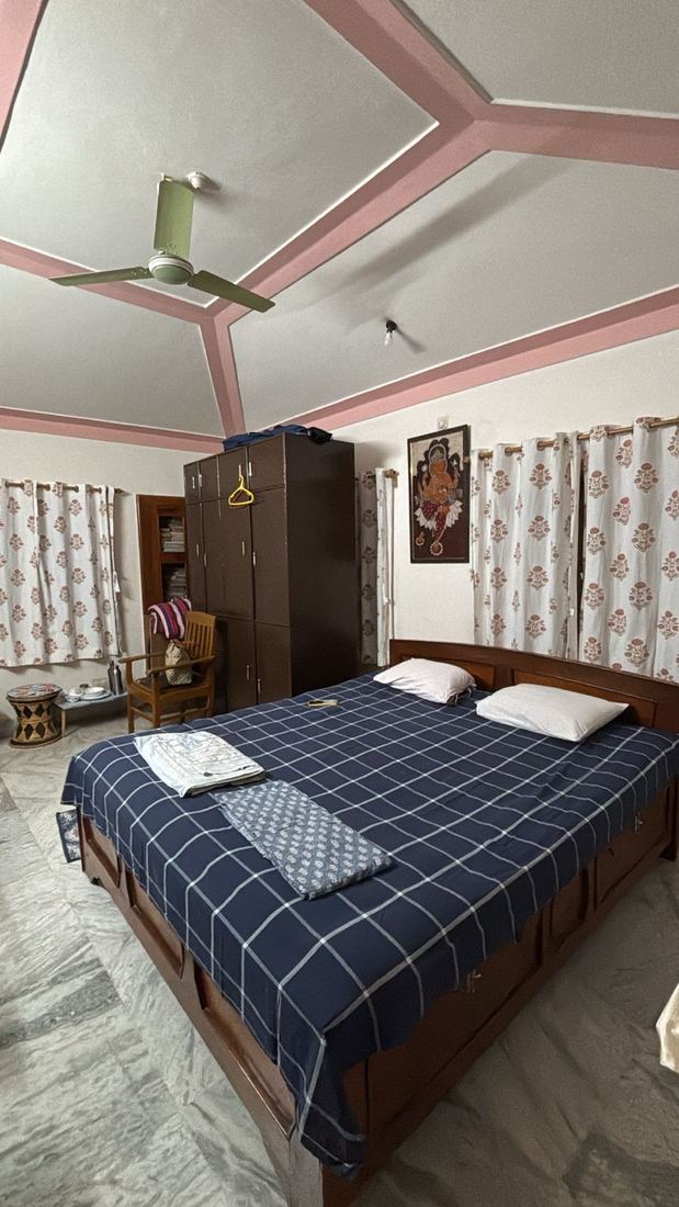 A bedroom with a pink-panelled ceiling, a green ceiling fan, blue checked bedspread on a wooden storage bed, a tall almirah, a framed pattachitra of Ganesha, and block-print curtains on two walls.