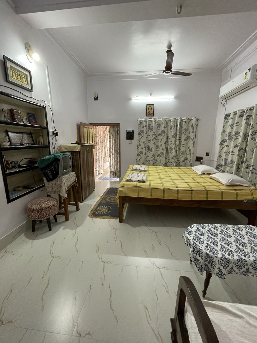 A large high-ceilinged room with a yellow checked bedspread on a wooden bed, ceiling fan, air conditioner, a shelf of books and curios, and an open door through to the balcony.