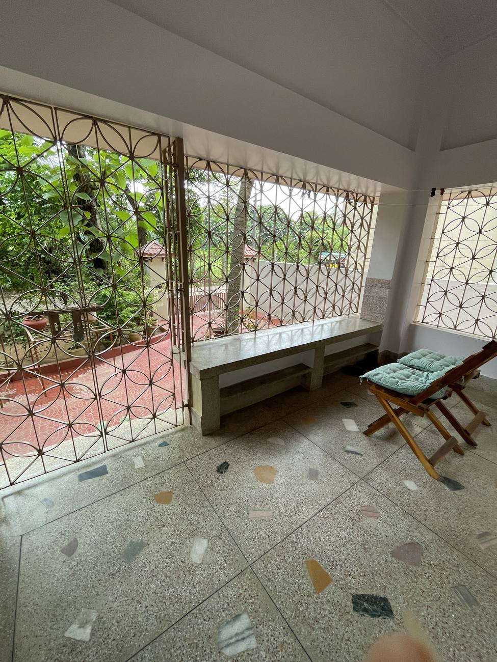 The verandah: terrazzo floor with coloured marble chips, a long concrete bench under a wall of four-petal iron grilles, a wooden folding chair with green cushions.